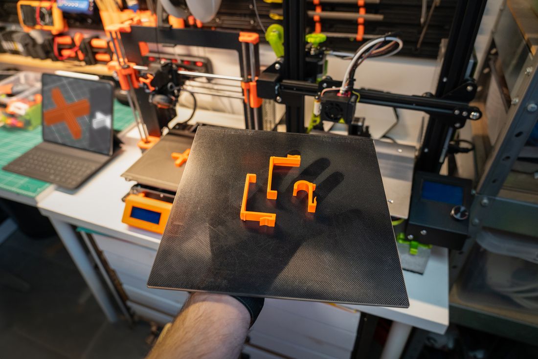 A maker holds up a print plate with orange printed brackets in front of a workshop full of 3D printers