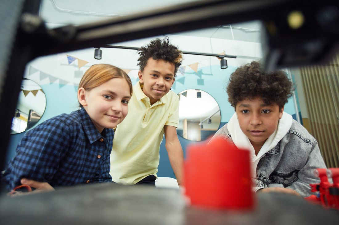 Three young people lean in to watch a red part being 3D printed