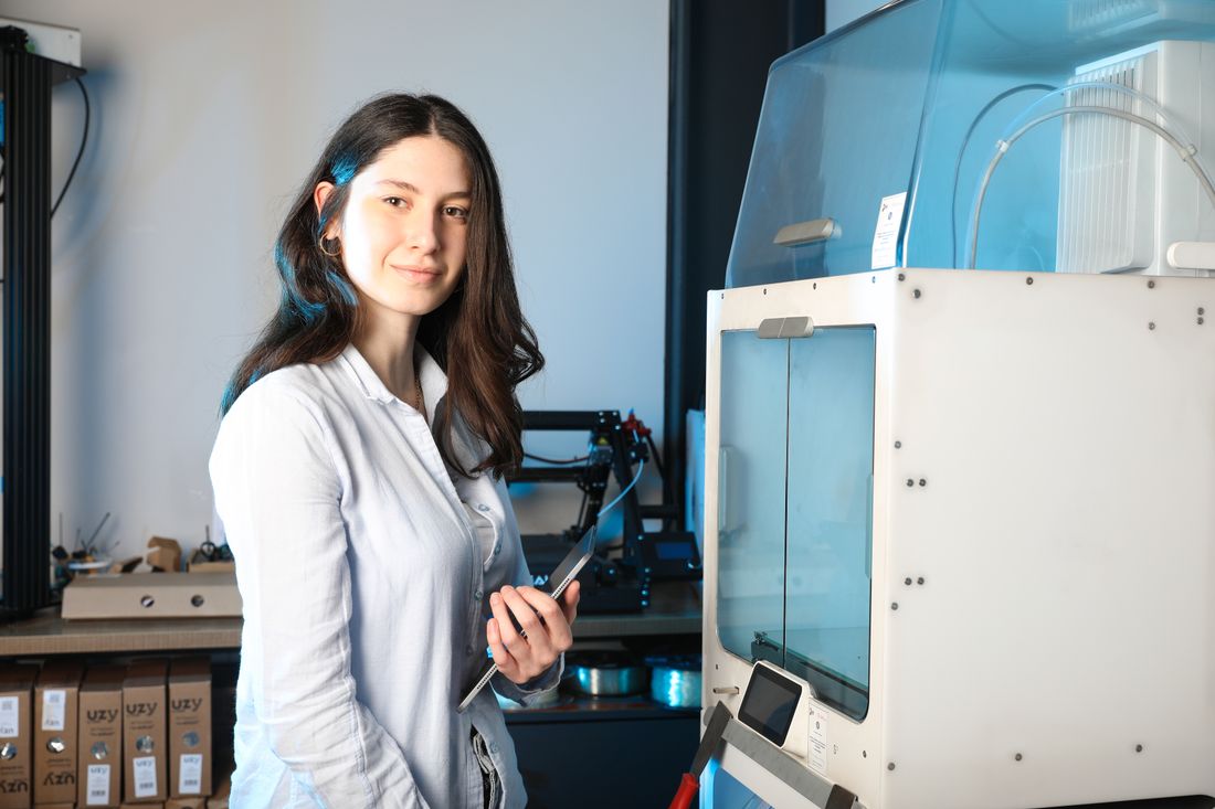 A woman holding a tablet stands beside an enclosed 3D printer in a workshop