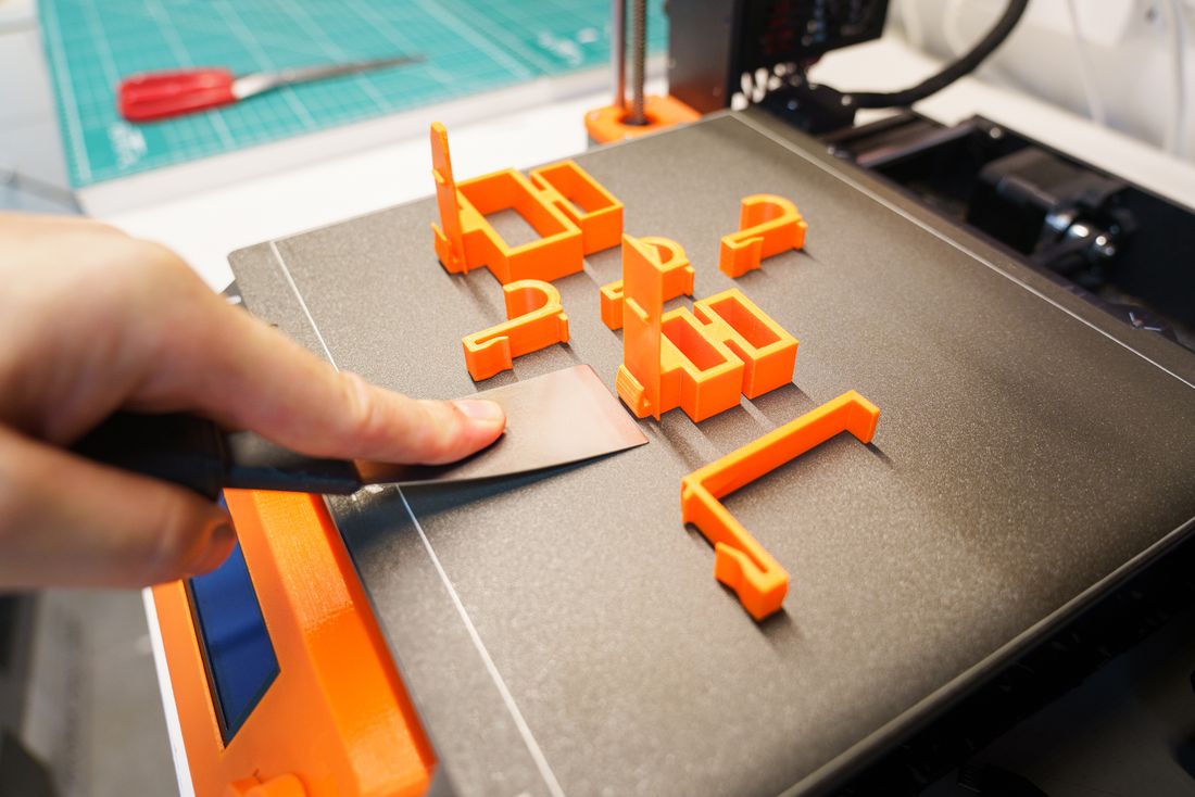 A hand uses a scraper to lift freshly printed orange parts off a 3D printer bed